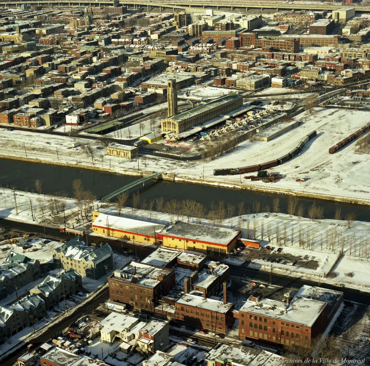 Vue oblique d’une ville de Saint‑Henri enneigée, bâtiment en premier plan, rues bordées de voitures enneigées.